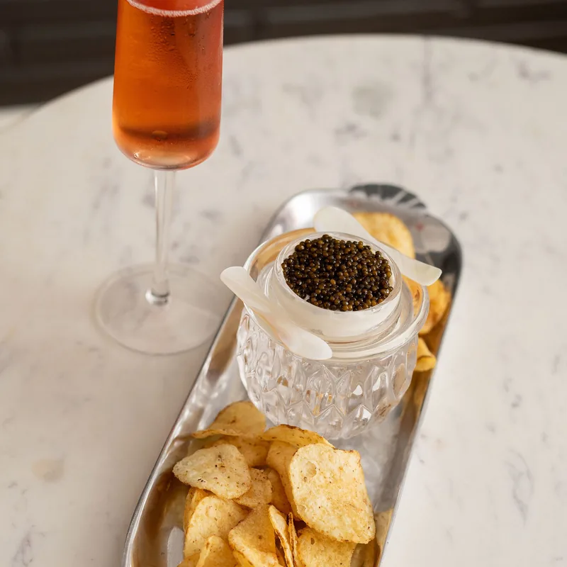 Caviar service in a cut-crystal jar on a silver tray with kettle chips, beside a coupe of sparkling rosé.