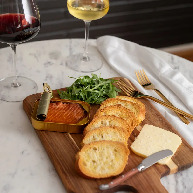 Glasses of red and white wine on a marble table beside a board of smoked trout, arugula and toasted baguette.