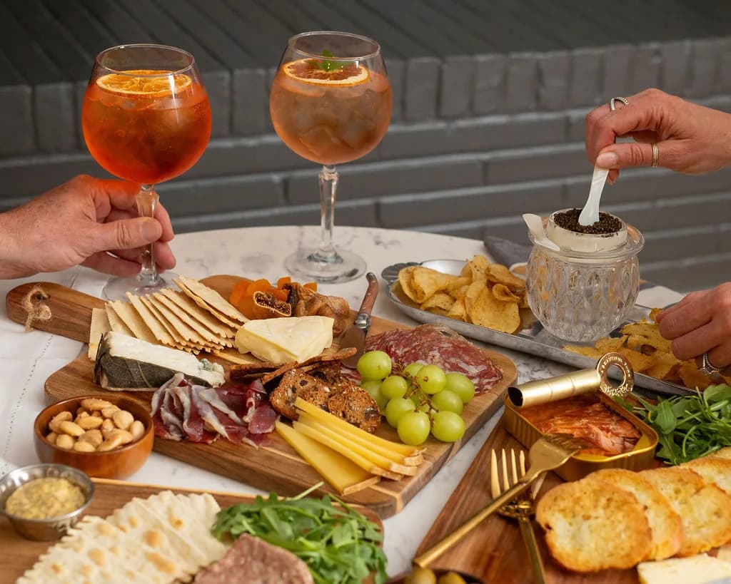 Two people sharing a spread across a marble table: a wooden board of crackers, brie, blue cheese, dried apricots and figs, salami, grapes and batons of hard cheese, with a bowl of marcona almonds, a tray of kettle chips and a tin of preserved fish alongside. One hand lifts a spritz garnished with dried orange; another spoons caviar from a cut-crystal jar with a mother-of-pearl spoon.