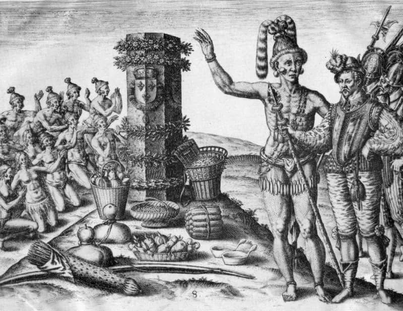 A 1591 engraving of Timucua people gathered around the stone column raised by French Huguenot colonists near the River May, now the St. Johns. The column, carved with the French royal fleur-de-lis and garlanded with foliage, is ringed with baskets of maize, fruit and game left as offerings; a Timucua leader gestures toward it while two Frenchmen in doublets look on.