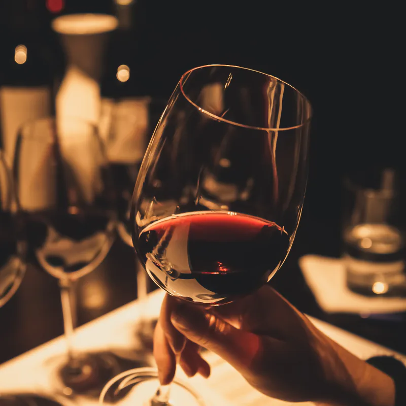 A hand raising a glass of red wine across a candlelit table, with bottles and other glasses blurred behind it.